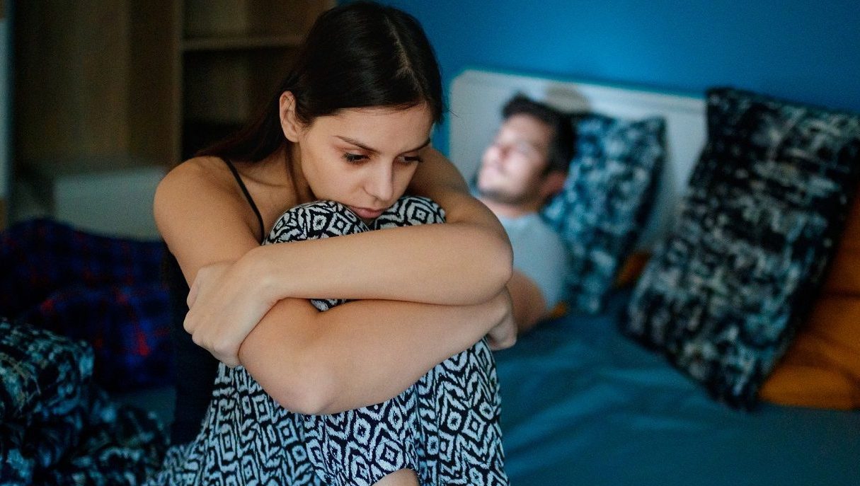 Sad Woman Sitting On Bed With Partner In Background