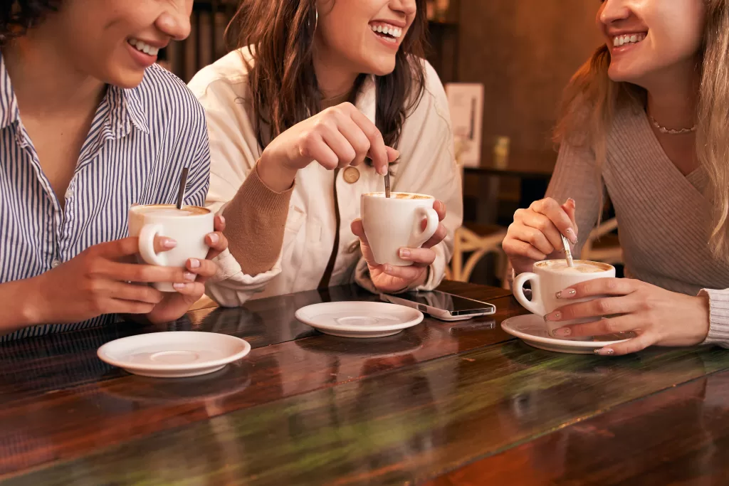 Group Of Three Unrecognizable Girl Friends Drinking Coffee Sitting In A Table Of A Cafe.