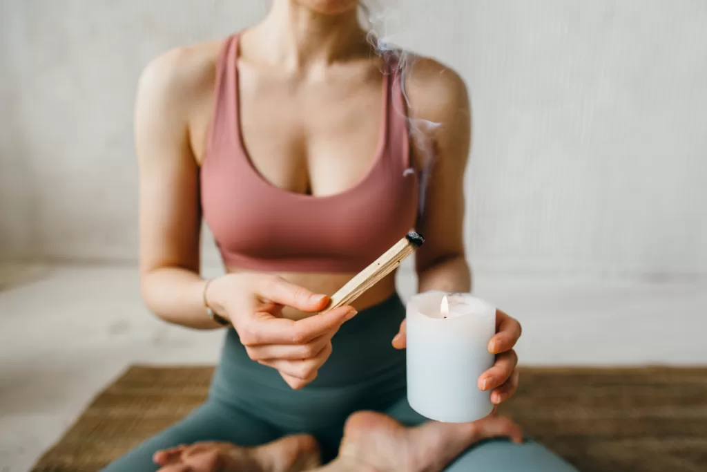 Young Woman Lights A Palo Santo Stick From A Candle Before Meditation. The Process Of The Ritual Of Fumigating A Room For Spiritual Practice.